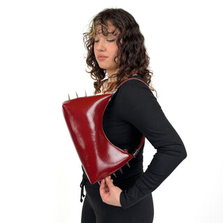 A person holding a black glossy vegan leather shoulder bag with silver spikes, designed to be worn across the shoulder or as a hand clutch.